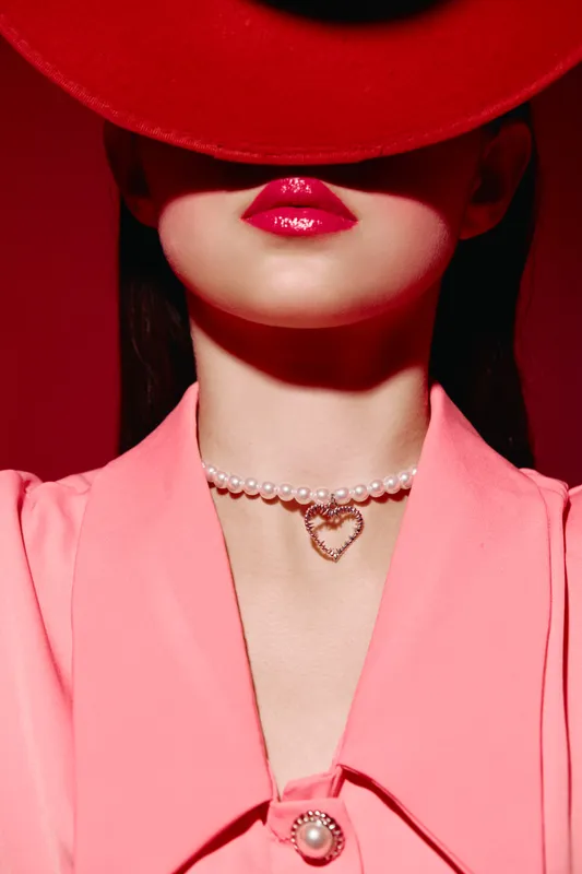 Close-up of a woman with glossy red lips wearing a bright red hat and pink blazer with pearl detail. Creative fashion portrait in bold colors showcasing beauty and modern style.