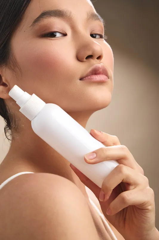 asian woman holds a spray bottle near her face in a skincare scene, exuding calm focus and selfcare as she prepares a beauty routine in a pristine studio setting.