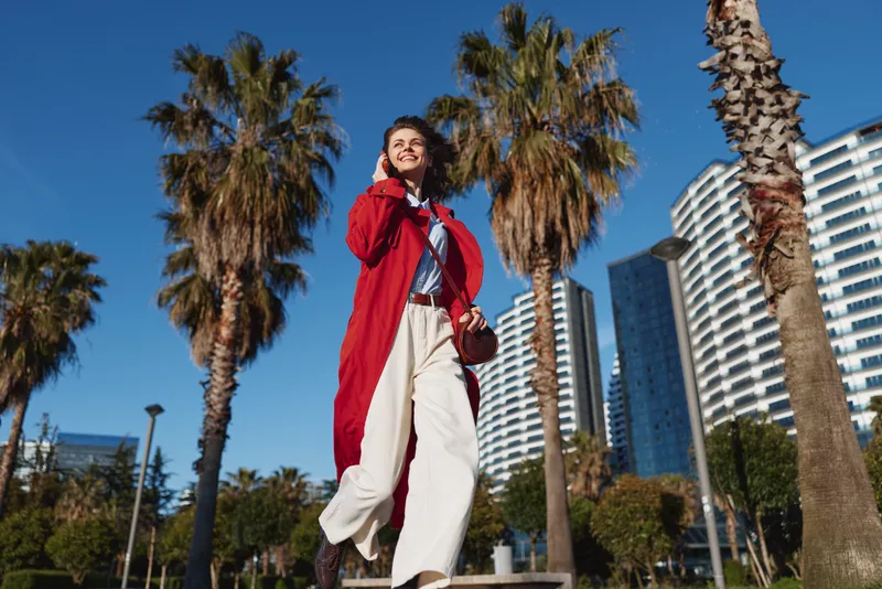 A young woman wearing a red coat and white trousers walks confidently along a sunny city street framed by palm trees and modern buildings.