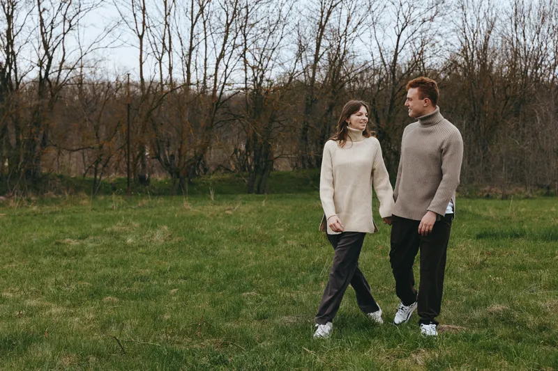 Young couple wearing cozy sweaters walks together across a green field in early spring while holding hands and enjoying a peaceful moment outdoors.