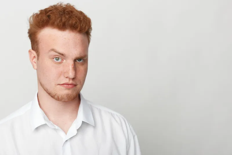 Portrait of a serious young man with red hair and freckles wearing a white shirt against a plain light background. Concept of confidence, youth, and natural appearance.