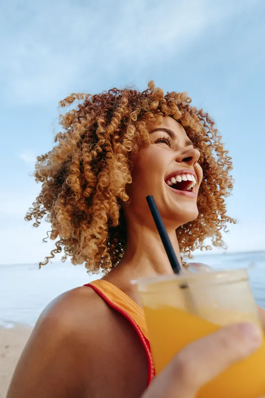 Happy woman with curly hair holding a cold drink with a straw outdoors. Bright sunny day with blue sky. Concept of summer, refreshment, and joy in nature.