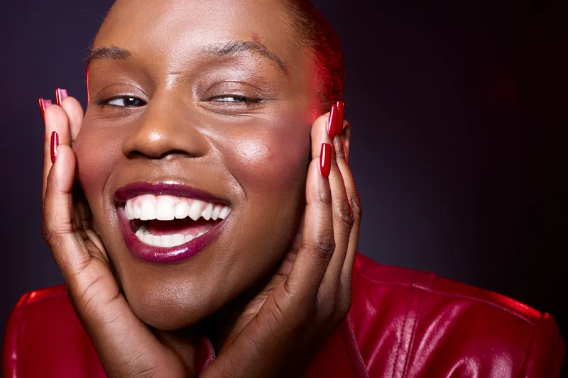 Close-up of joyful black woman with dark skin smiling broadly, wearing red leather jacket and dark red lipstick. Concept for happiness, beauty, and self-confidence in studio lighting.