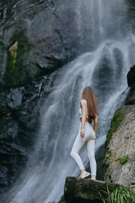 Woman standing near a waterfall in white sportswear, long hair flowing, surrounded by lush greenery and rocks, showcasing nature and active lifestyle in misty mountain setting.
