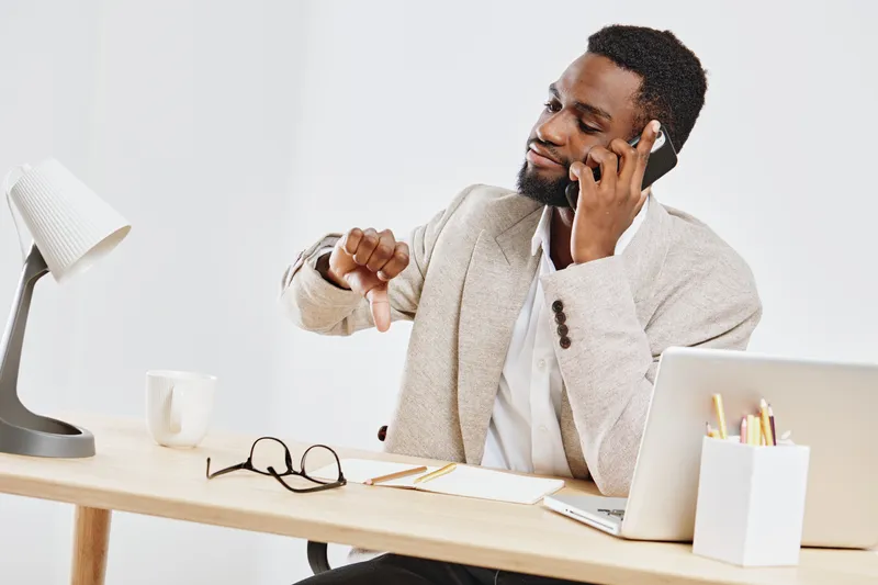 Professional man in beige blazer talking on mobile phone while working at desk with laptop, glasses, notebooks, and lamp. Modern office workspace, business communication, and remote work concept.