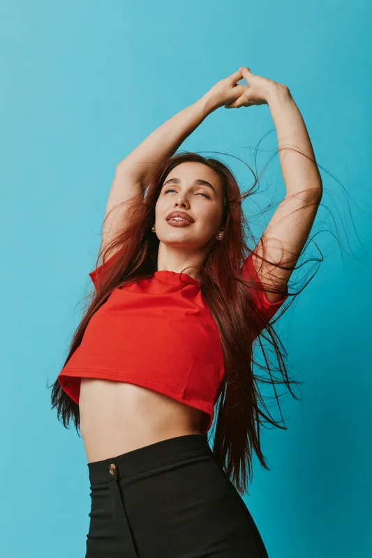 Young woman in red crop top stretching with eyes closed and hair flowing against blue background, showing relaxation, freedom, and natural beauty in casual fashion style.