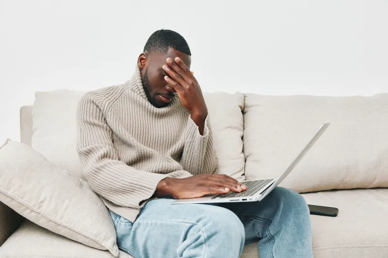 Young Black man stressed while working on laptop at home in cozy sweater, conveying frustration and uncertainty in a modern setting