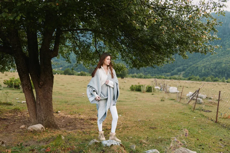 A woman enjoys an outdoor lifestyle beside a tree in a rural field, wearing a light cardigan and jeans, embracing nature and fresh air on a sunny day.
