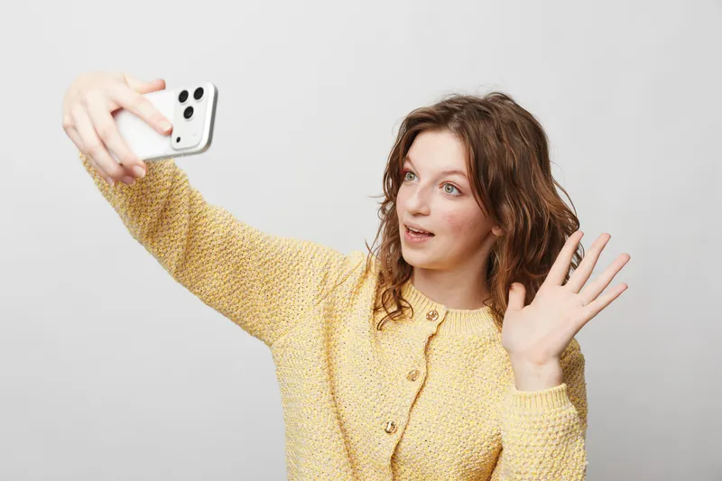 Young woman with curly hair wearing yellow sweater takes selfie with smartphone, smiling and waving hand. Bright studio background, concept of communication, social media, and technology use.