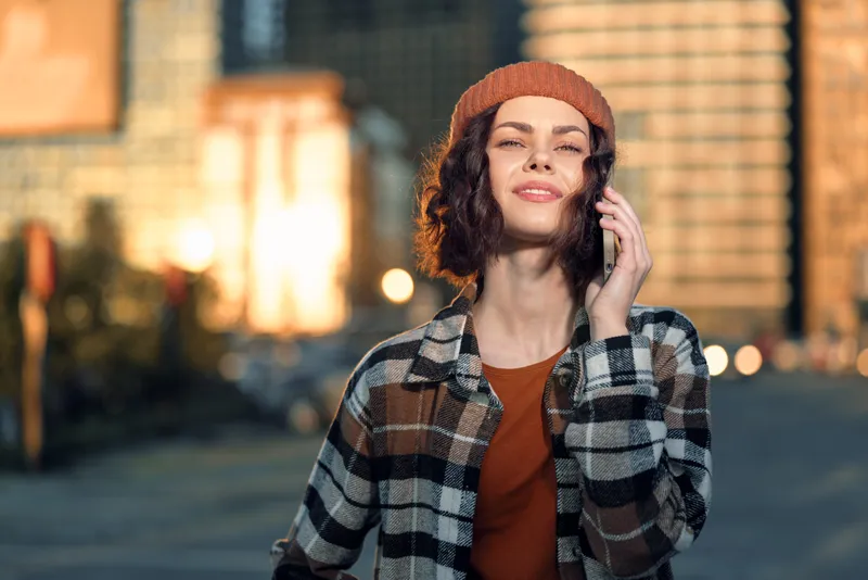 woman phone urban golden hour candid lifestyle beanie portrait: Young woman smiling on phone in city street at golden hour glow, casual fashion and curly hair, authenticity, mindful living and