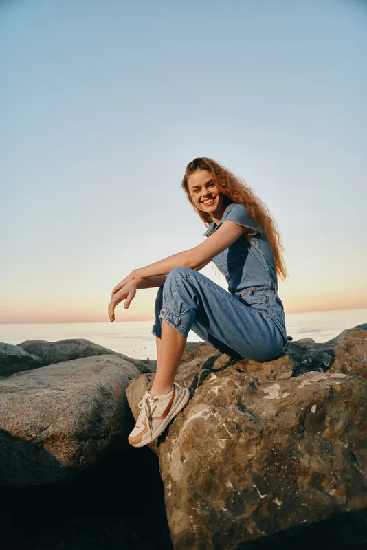 Lifestyle woman in modern boho-western denim street style sitting on rocks by the sea during sunset, film color tones creating a warm and relaxed atmosphere.