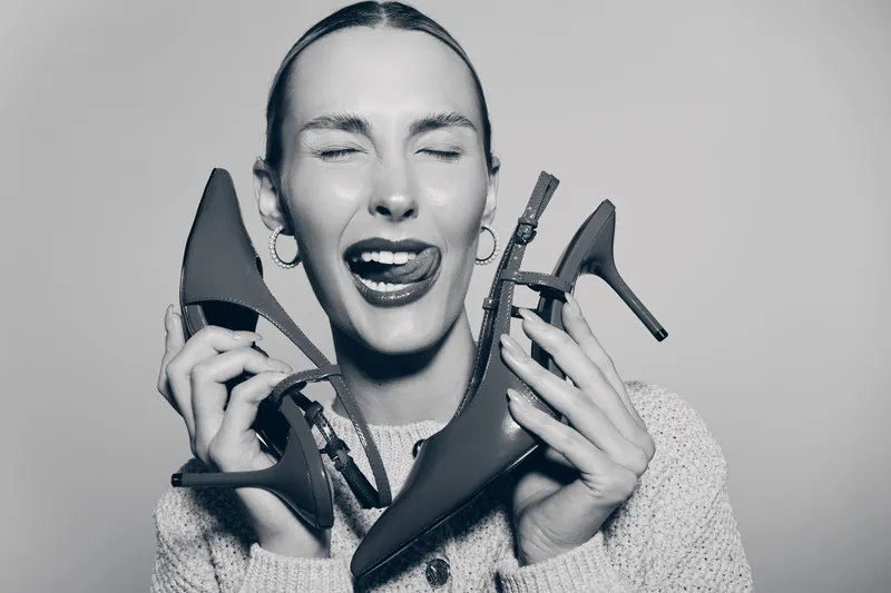 A happy white woman with pale skin and slicked back hair holds two high heel shoes near her face, eyes closed and tongue out. Concept of fashion, joy and personal style.