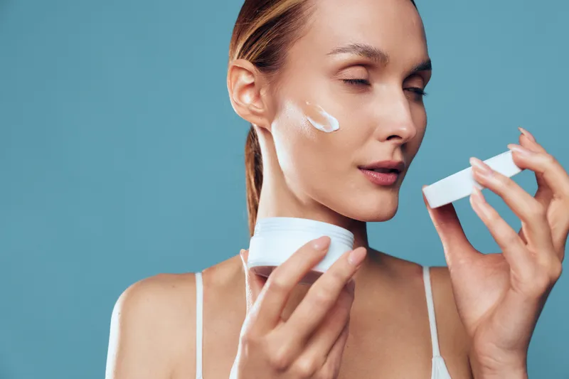 A young woman applies white cream on her cheek with closed eyes. She holds the cream container in her hand against a plain blue background. Concept for skincare and beauty routine.