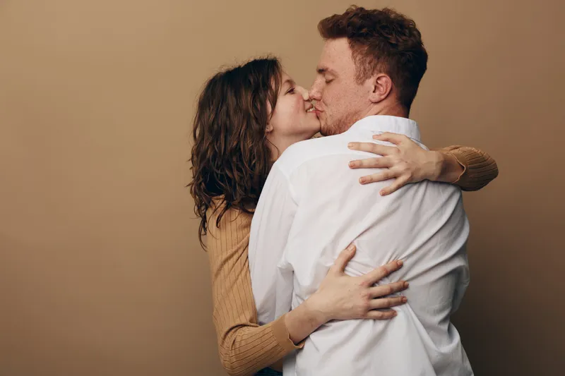 Young couple embraces and kisses with closed eyes on beige background. Intimate moment of love and affection between man and woman in casual clothing. Concept of romance and connection.