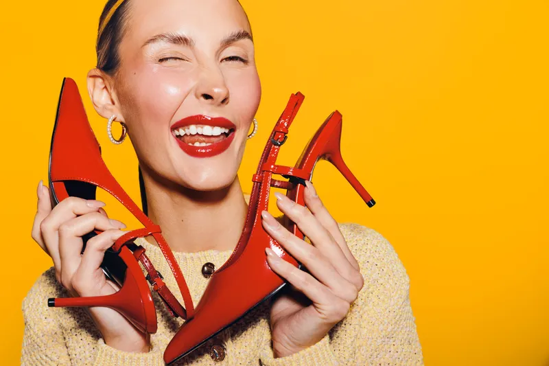 Happy woman with red lipstick holds red high heel shoes near face, winking and smiling on bright yellow background. Concept of fashion, style, beauty, and joyful emotions.