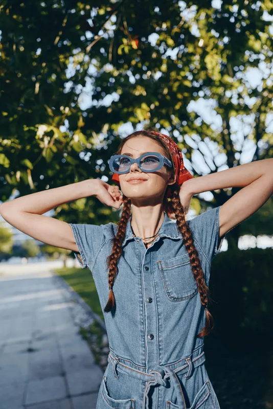 Lifestyle woman in modern boho-western denim street style enjoys sunny day outdoors wearing sunglasses and red bandana with braids against leafy background.