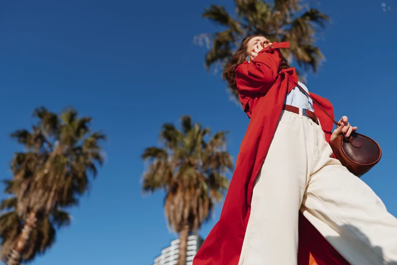 A young woman in a bright red coat strides confidently outdoors against a clear blue sky, white wide leg pants fluttering, palm trees rising behind.