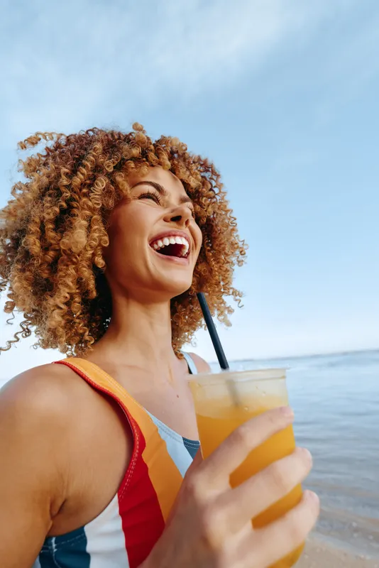 One person, young woman with curly hair laughs and holds an orange drink on a sunny beach in a striped swimsuit, conveying carefree summertime mood and festive vibe.