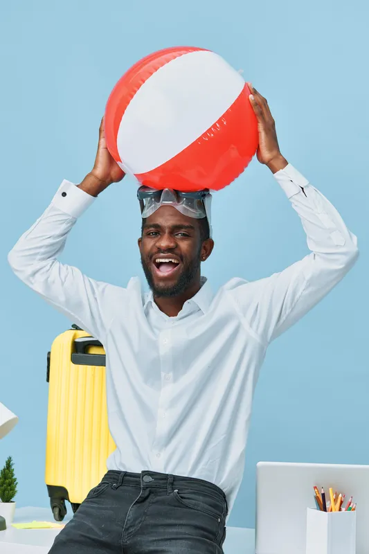 Joyful young man with dark skin and a wide smile holds a colorful beach ball above his head, wearing a white shirt and sunglasses, standing against a light blue background. People lifestyle concept, s