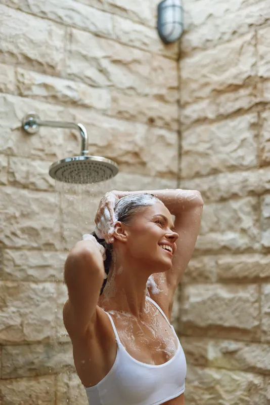 woman showering outdoors with wet hair, smiling and relaxed under rain showerhead against stone wall background, natural light and casual attire