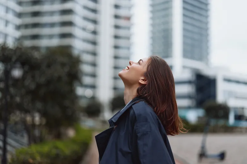 Young woman with brown hair wearing dark coat looking up in the city street. Urban lifestyle concept with modern buildings and natural light.