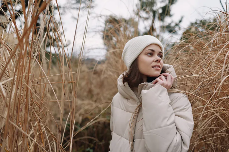 Winter outdoor portrait of a woman in a beige puffer jacket and a knit beanie among tall grasses, conveying calm weather and seasonal mood.