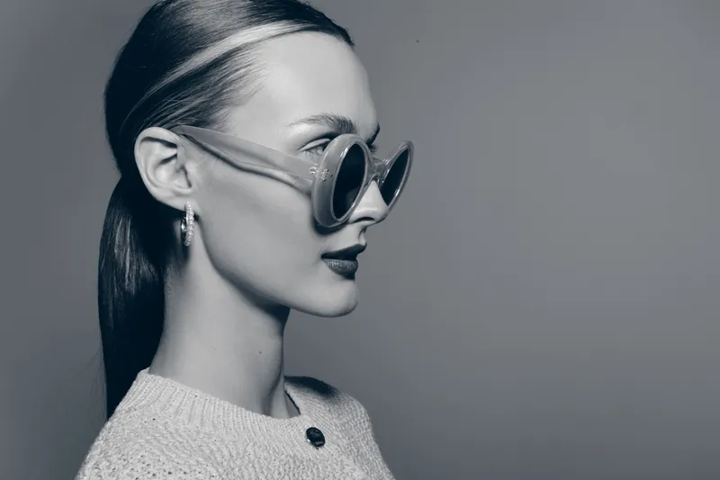 Profile of a pale skinned Caucasian woman with dark hair in ponytail wearing round sunglasses and a knitted sweater. Minimalist fashion concept with a stylish and elegant mood.