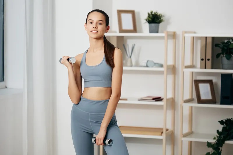 Fitness woman in athletic wear lifts dumbbells in a bright home gym, training pose with calm expression, minimalist shelves and plants in the background.
