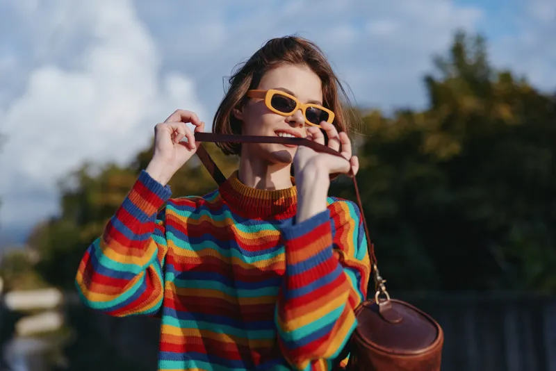 Woman in colorful striped sweater and yellow sunglasses smiles outdoors while playfully biting a leather bag strap, casual fashion portrait with sunny sky and trees.