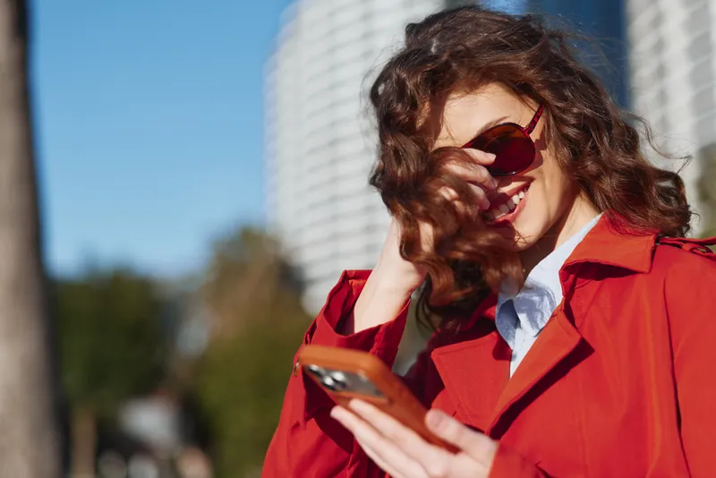 Young woman in a red coat laughs while looking at her phone in bright sun with a city backdrop, a carefree street moment captured with wind in her hair and bold color