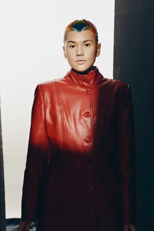 Young woman with colorful short hair wearing a stylish red leather coat standing confidently against a minimalistic background, modern fashion look.