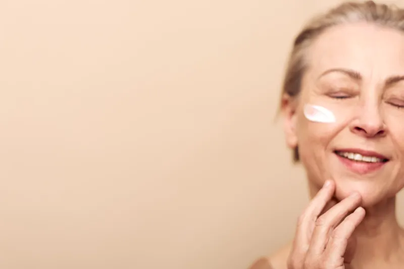 Smiling mature woman with closed eyes applying face cream on cheek. Concept of skincare, self-care, aging beauty, wellness, and natural healthy skin in soft warm light studio environment.