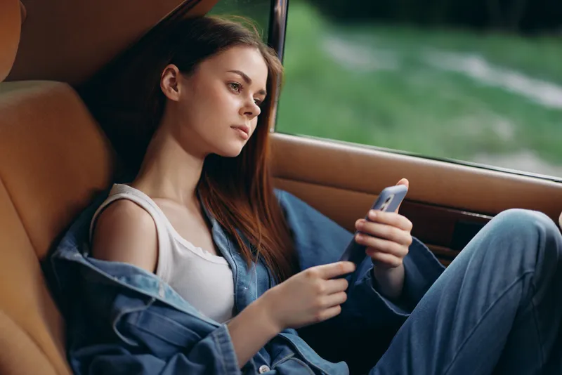 Thoughtful young woman in denim outfit, relaxing in a car while using smartphone, set against a green outdoor background, showcasing calm emotions.