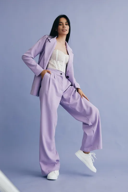 Young woman wearing a stylish lilac pantsuit and white sneakers posing confidently against a soft blue background. Modern, fashionable, poised, casual chic.
