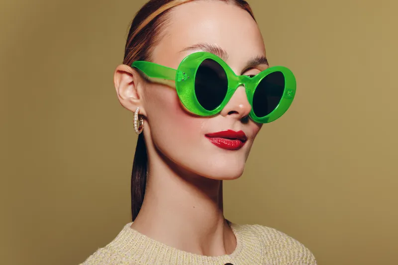 Young woman with green sunglasses and red lipstick poses against beige background. Fashion portrait with stylish accessories and confident expression. Concept of modern style and beauty.