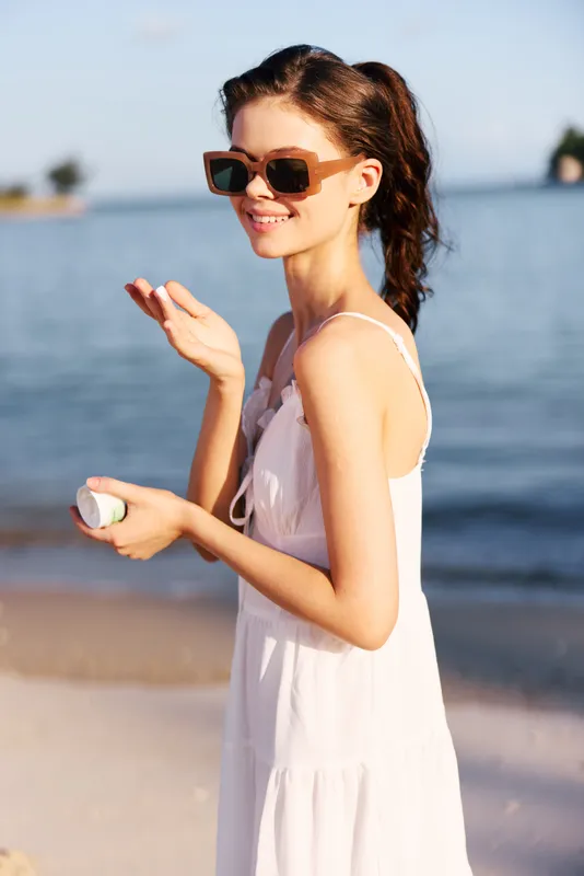 Smiling woman wearing sunglasses and white dress applies sunscreen near the beach on a sunny summer day, enjoying warmth and outdoor relaxation.