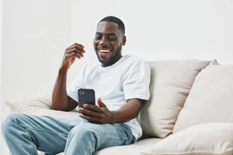 Happy young African man sitting on a beige sofa, enjoying a video call on his smartphone with a joyful expression, relaxed atmosphere and soft lighting