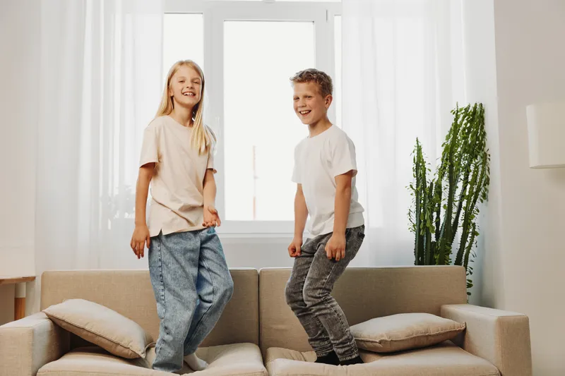 Happy children jumping on sofa in bright living room with natural light and casual clothes. Concept of childhood fun, energy, and home leisure activities indoors.