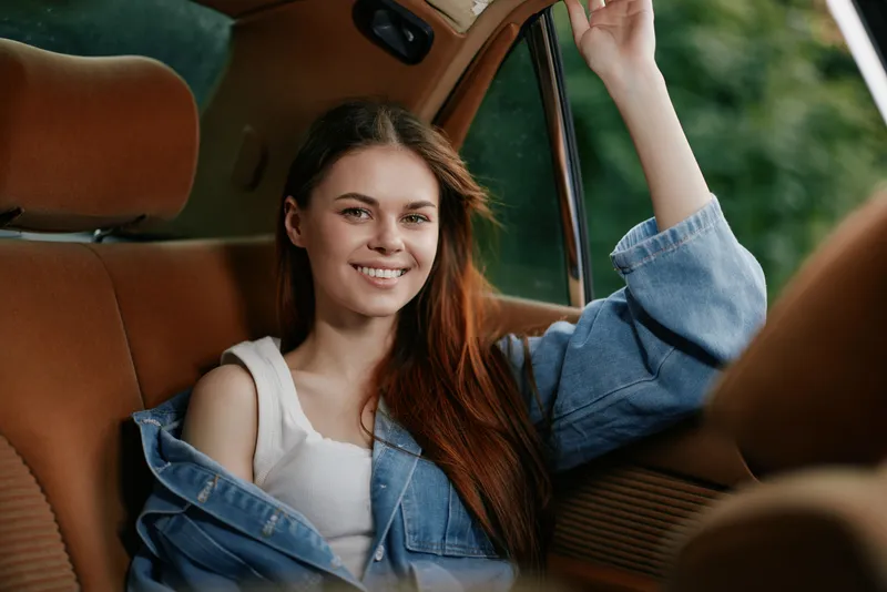 Young woman in casual attire smiling in a vintage car, showcasing confidence and relaxation against a lush green background.