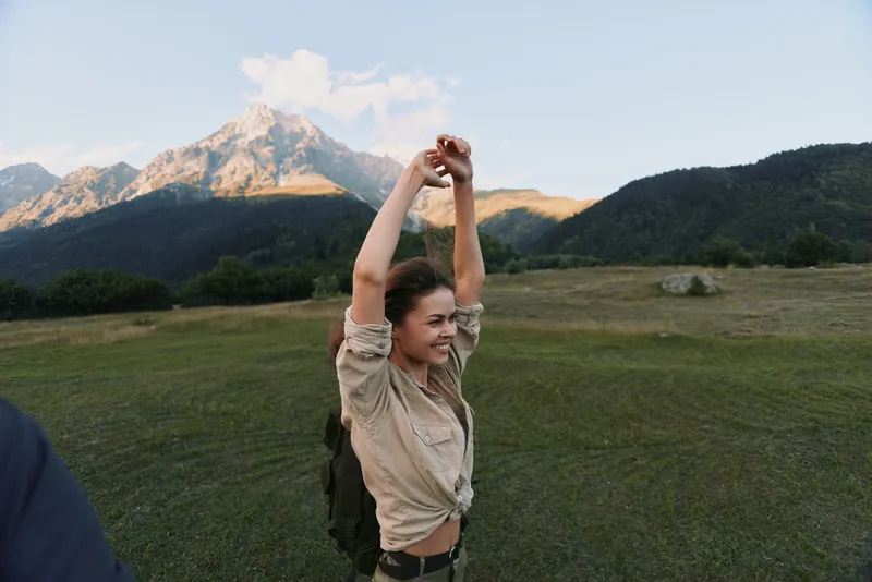 A joyful woman outdoors in hiking attire stretches her arms in a grassy field, with towering mountains in the background, conveying freedom and outdoor adventure.