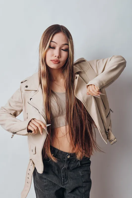 Young woman with long hair wearing beige jacket and crop top poses against light background. Fashionable model in casual outfit with confident expression and modern style in studio.