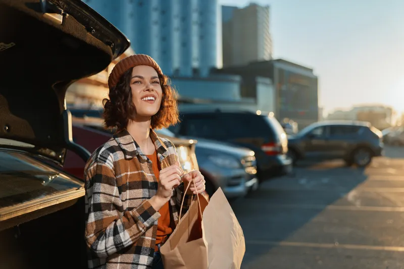 Woman by car in parking lot holding a paper shopping bag, smile and sunlight at golden hour glow. Candid lifestyle portrait with authenticity, mindful living and emotional storytelling vibe.