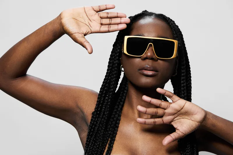 Young woman with long braids wearing oversized sunglasses, showcasing elegant poses against a minimalist gray background, exuding confidence and style