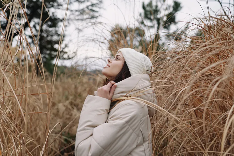 Winter portrait of a woman in a beige coat and beanie, standing among tall grass, gazing upward with a serene and reflective mood outdoors.