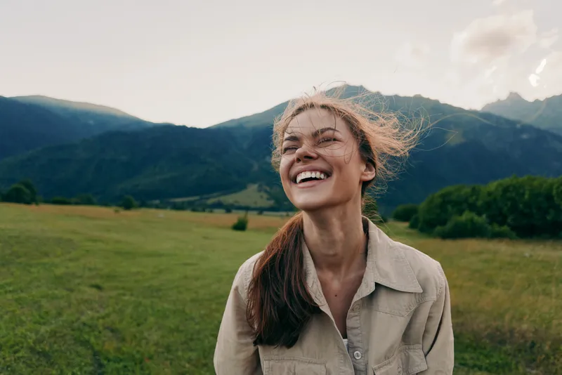 Young woman laughing outdoors in a sunlit valley with rolling green fields and distant mountains, wearing a beige shirt and enjoying the fresh breeze