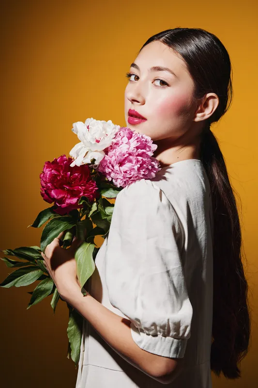 Young woman with glossy dark hair and red lipstick holds bright peony flowers against yellow background. Concept of beauty, creative fashion, floral elegance and softness.