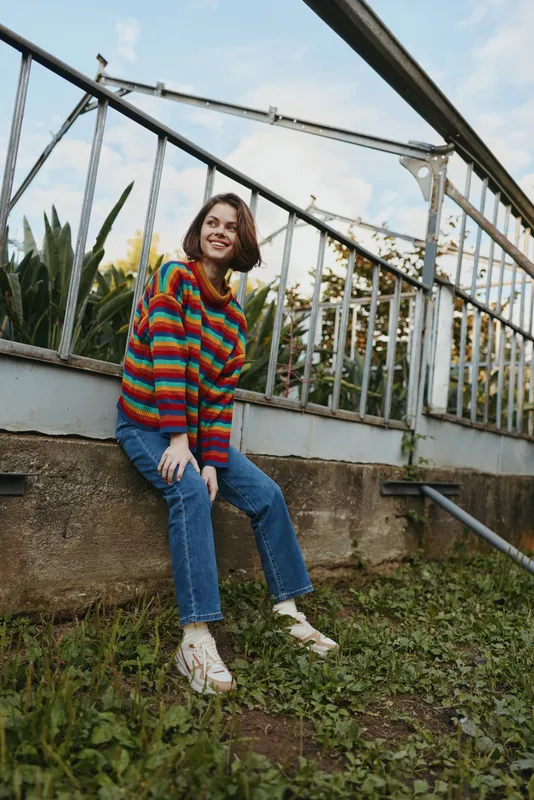 Young woman in a colorful striped sweater sitting on a low concrete wall in an urban garden, smiling and leaning forward in a casual outfit with blue jeans and white sneakers.