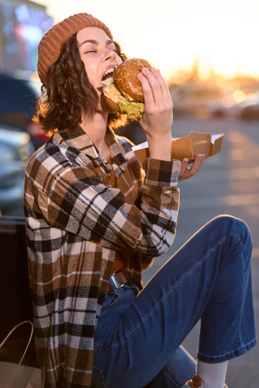 woman eating burger at golden hour, candid lifestyle urban scene with joyful smile and authenticity, emotional storytelling and mindful living captured in warm golden hour glow.