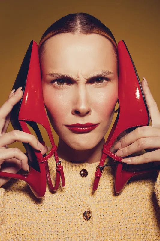 Young woman with red lipstick holds red high heels near face, making a funny expression. Studio portrait with warm lighting and yellow background. Concept of fashion and emotions.