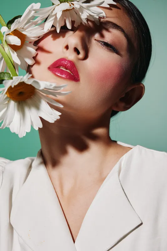 Close-up portrait of a creative beauty model with bold glossy red lips and soft blush. White flowers frame her face against a turquoise background. Fashion concept with artistic makeup.